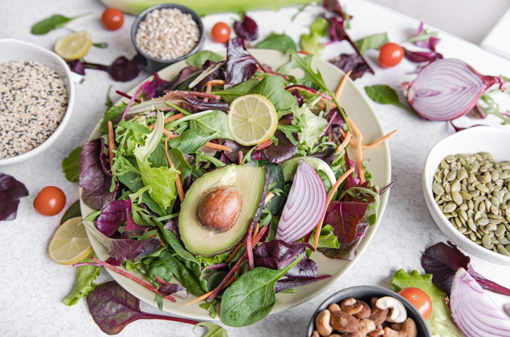 Osoba trzyma w dłoniach różową miskę wypełnioną zieloną sałatką z brokułami, pistacjami i kawałkami tofu, na tle kwiatów magnolii, reprezentując posiłek z wysokobiałkowej diety wegetariańskiej.