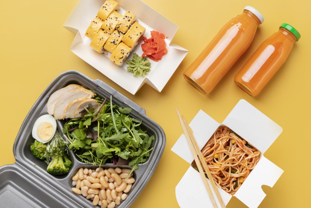 Zestaw różnorodnych posiłków na wynos na żółtym tle, obejmujący pojemnik z kurczakiem, warzywami i jajkiem, pudełko z sushi oraz makaron, prezentujący jak może wyglądać dieta pudełkowa bez glutenu i laktozy.