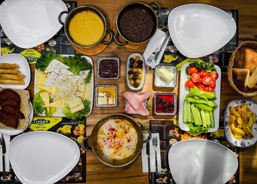 Widok z góry na stół zastawiony licznymi potrawami śniadaniowymi, takimi jak szakszuka, sery, świeże warzywa i dipy, prezentujący catering z dostawą do domu.