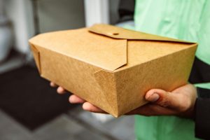 Kurier dostarczający catering wegetariański trzyma w dłoniach brązowe, papierowe pudełko z posiłkiem.